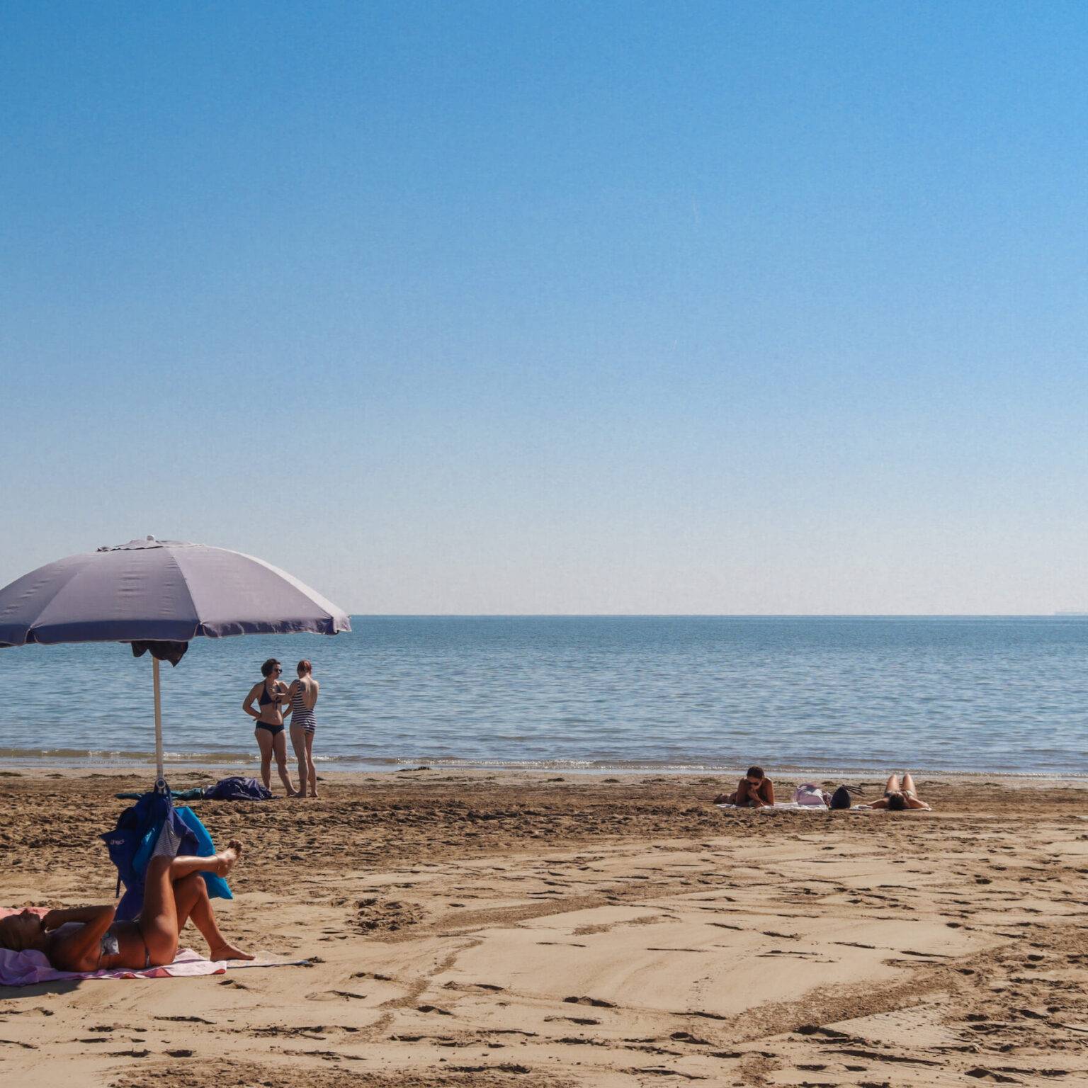 Der lange Sandstrand und das Meer beim Strandbad Bluemoon. Vorne links sitzt eine Person unter einem Sonnenschirm.