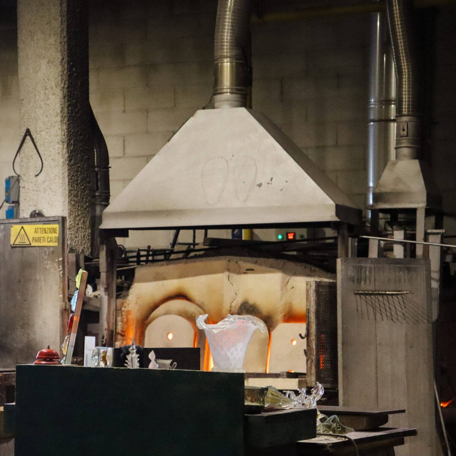 Der Ofen einer Glasbläserei im Hintergrund. Davor eine Auswahl verschiedener Stücke aus Glas.