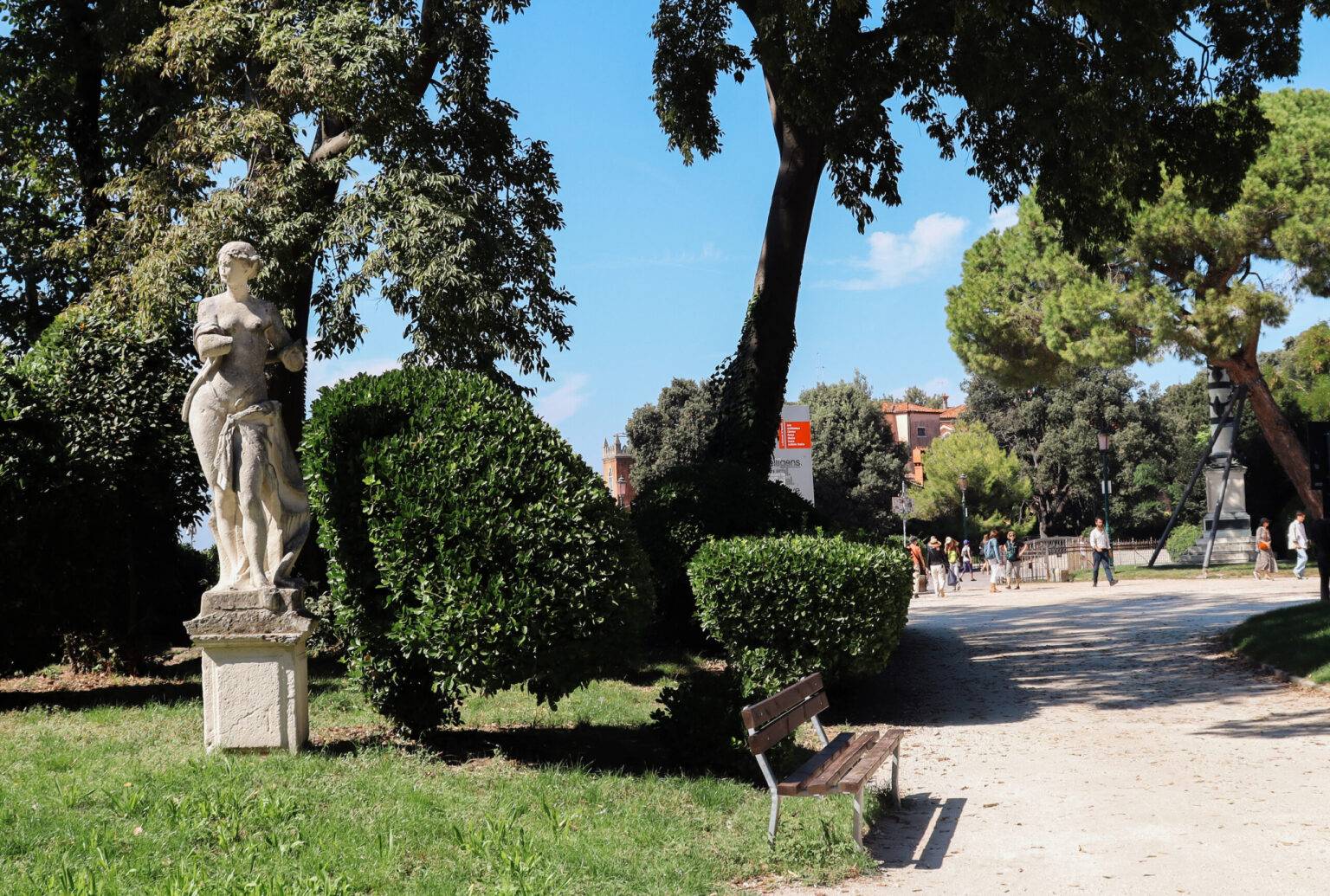 Ein Kiesweg und links grüne Bäume, Hecken, Wiese und eine Steinstatue im Biennale-Garten.