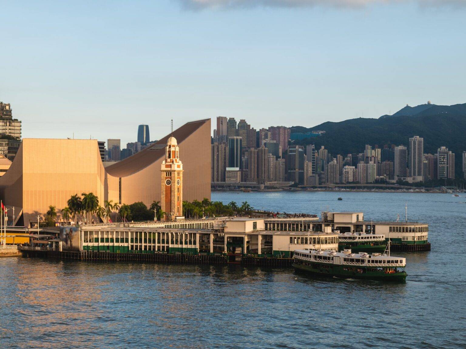 Perspektive vom Wasser auf das Ufer von Tsim Sha Tsui in Hongkong bei Sonnenuntergang mit Fähren und einem Kirchturm.