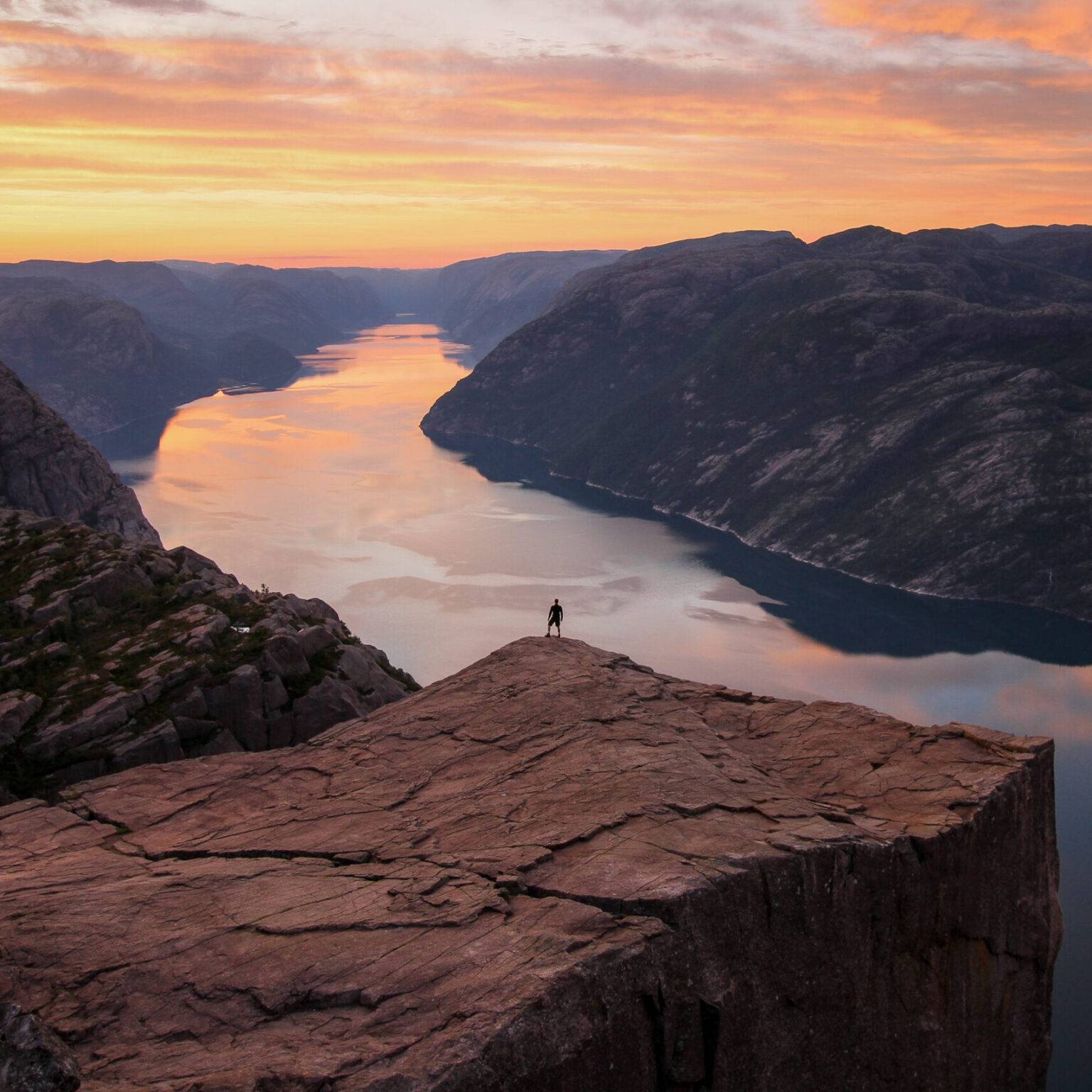 Ein Mann steht bei Sonnenaufgang auf dem Felsplateau Preikestolen. Zwischen den Felsen links und rechts fließt ein breiter Fluss.