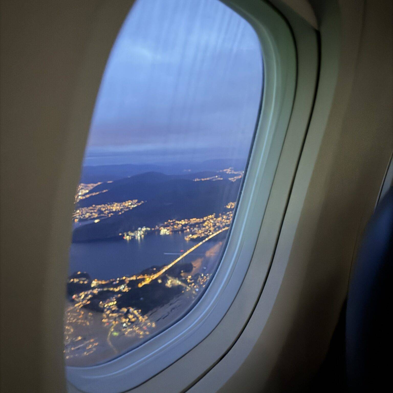Blick aus einem Flugzeugfenster auf das beleuchtete Bergen in der Dämmerung.