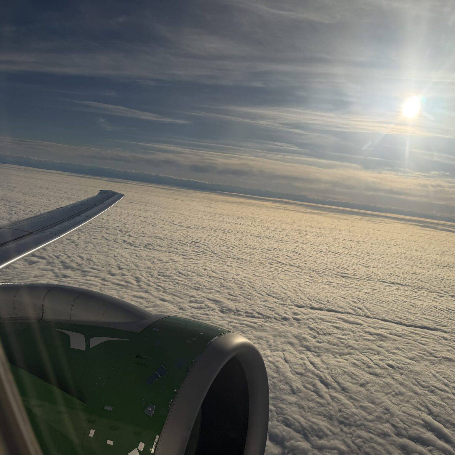 Blick aus dem Flugzeugfenster auf eine Wolkendecke und die Sonne am Himmel.