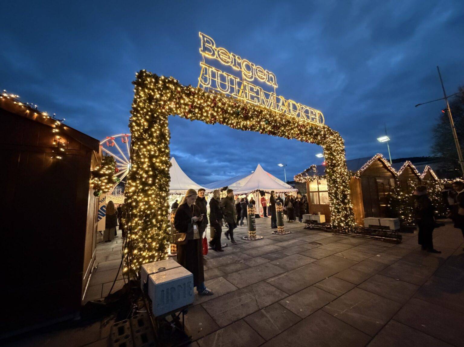 Beleuchteter Eingang zum Bergener Weihnachtsmarkt, dem Julemarked, mit Ständen und Besuchern unter einem dunklen Himmel.