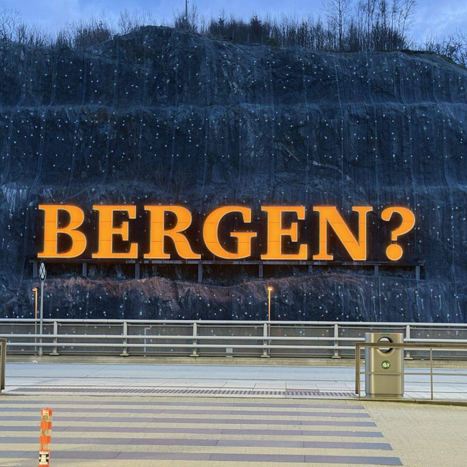 Große orangefarbene Schrift mit der Aufschrift "BERGEN?" vor einer felsigen, bewachsenen Wand, im Vordergrund ein Fußgängerübergang mit Zebrastreifen.