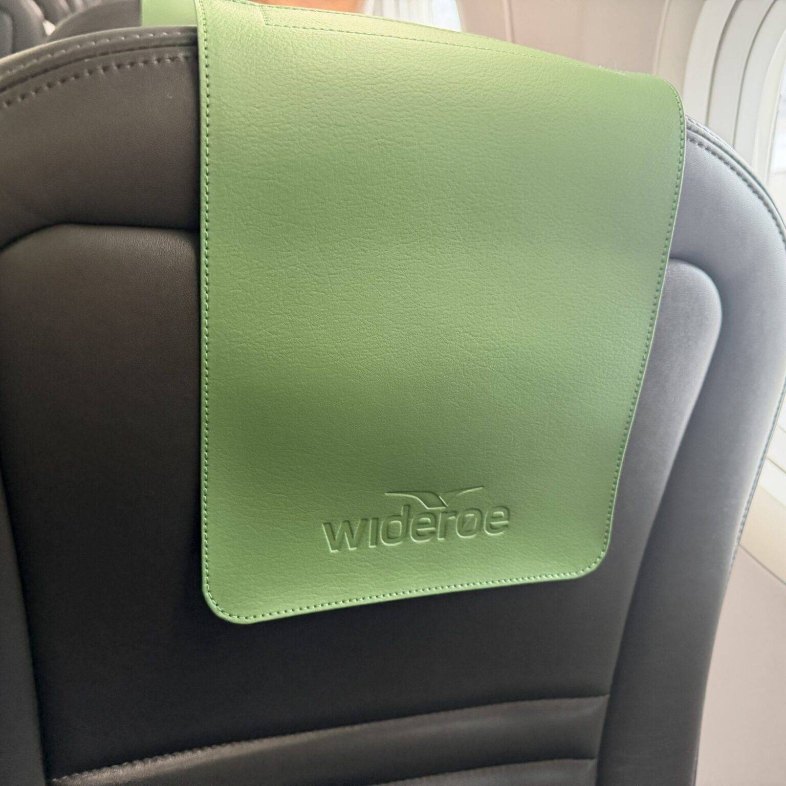 Grüner Bezug an einem schwarzen Flugzeugsitz, mit dem Logo von "wideroe" sichtbar.