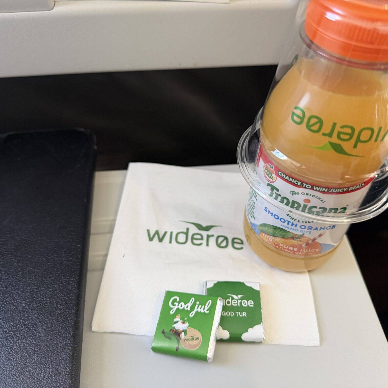 Flasche mit Orangensaft, Serviette mit "widerøe" und zwei Schokoladenstückchen auf einem Tisch, Hochformat.