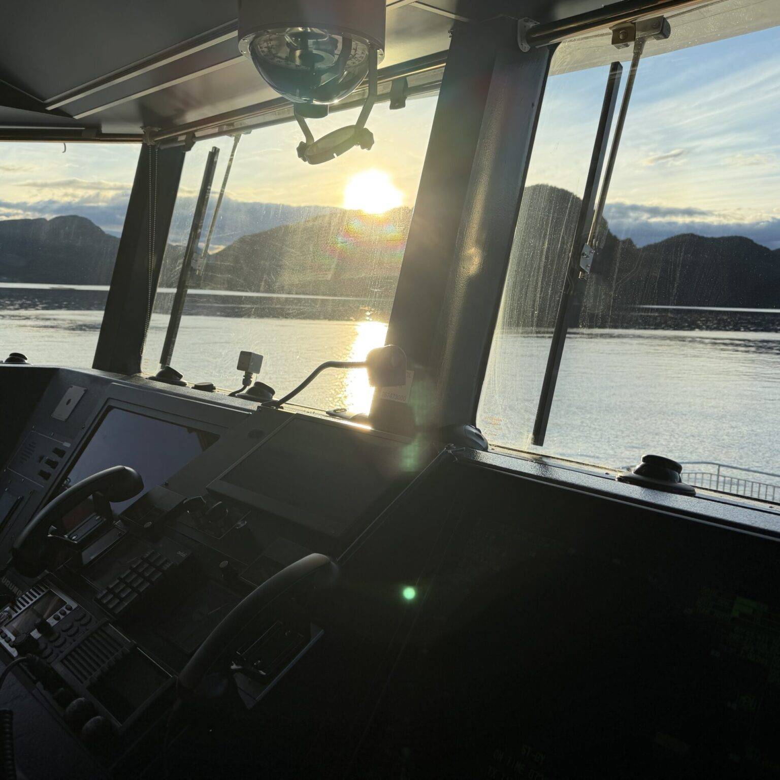 Blick aus dem Steuerstand eines Bootes auf einen Sonnenuntergang über dem Wasser und den umliegenden Bergen.