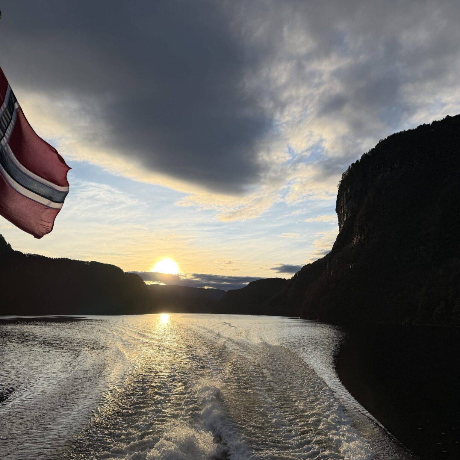 Norwegische Flagge weht vor einer Fjordlandschaft bei Sonnenuntergang, mit reflektierendem Wasser und bewölktem Himmel.