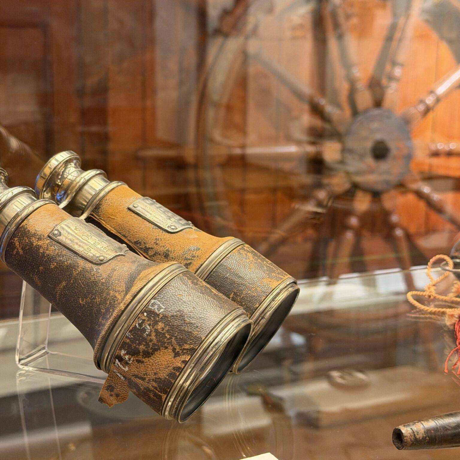 Zwei verrostete Ferngläser stehen in einer Vitrine, im Hintergrund ist ein historisches Steuerrad sichtbar.