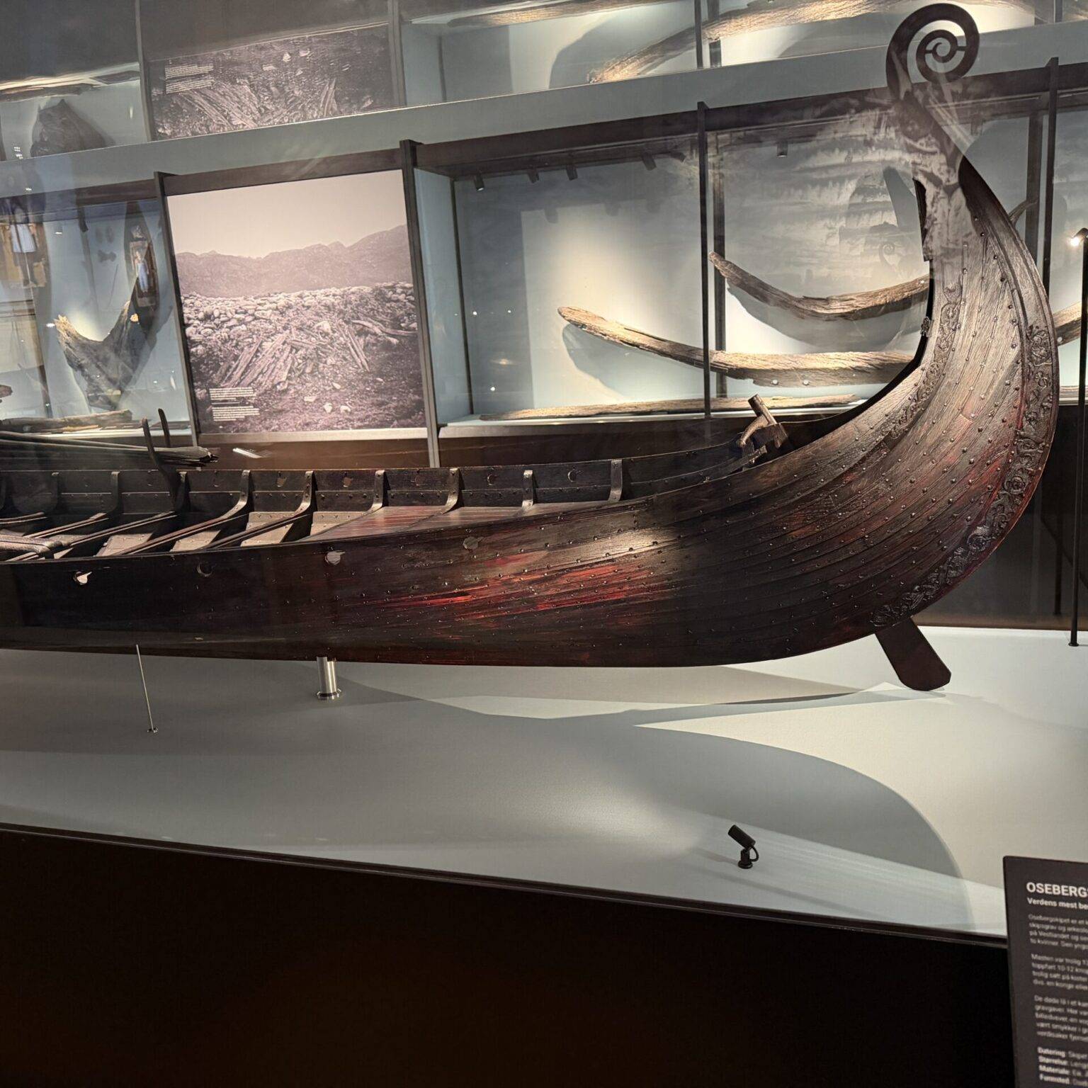 Ein historisches Wikingerschiff in einem Museum, ausgestellt hinter Glas mit weiteren Booten im Hintergrund.