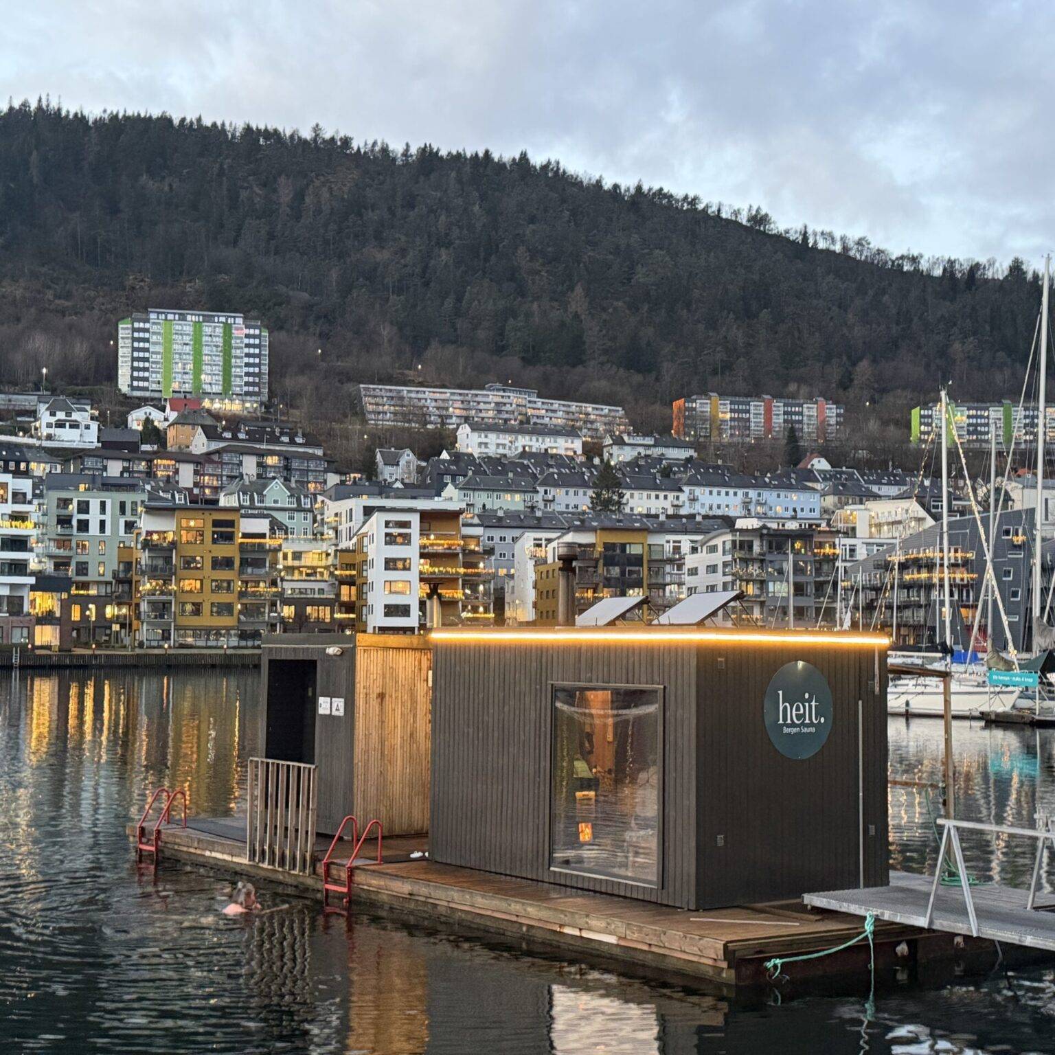 Modernes, beleuchtetes Gebäude auf dem Wasser im Hafen von Bergen, im Hintergrund ein Wohnviertel am Fuß eines Berges.