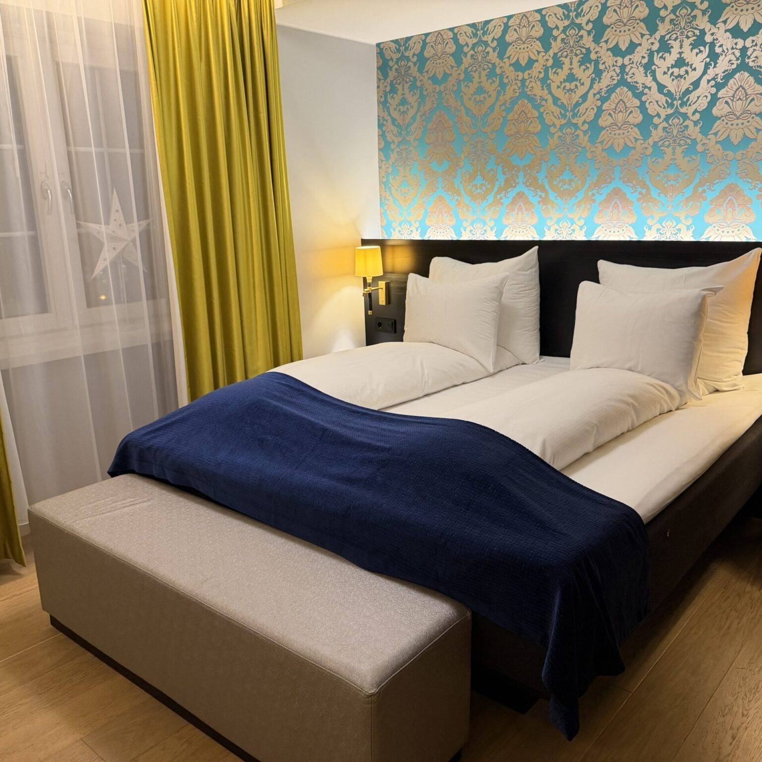 Stilvolles Hotelzimmer mit einem Doppelbett, blauer Decke, gelben Vorhängen und einer Sitzbank am Fußende.