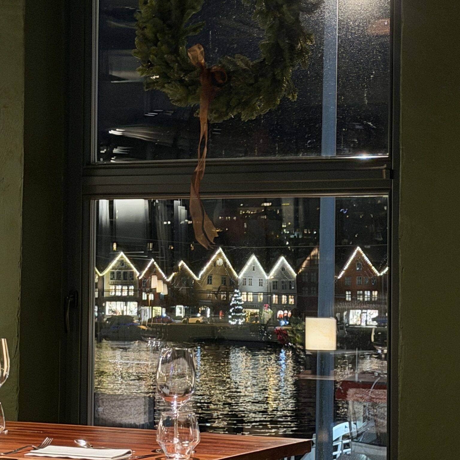 Fenster mit einem Kranz, Blick auf beleuchtete Häuser am Wasser und einem Tisch mit Glas und Besteck im Vordergrund.