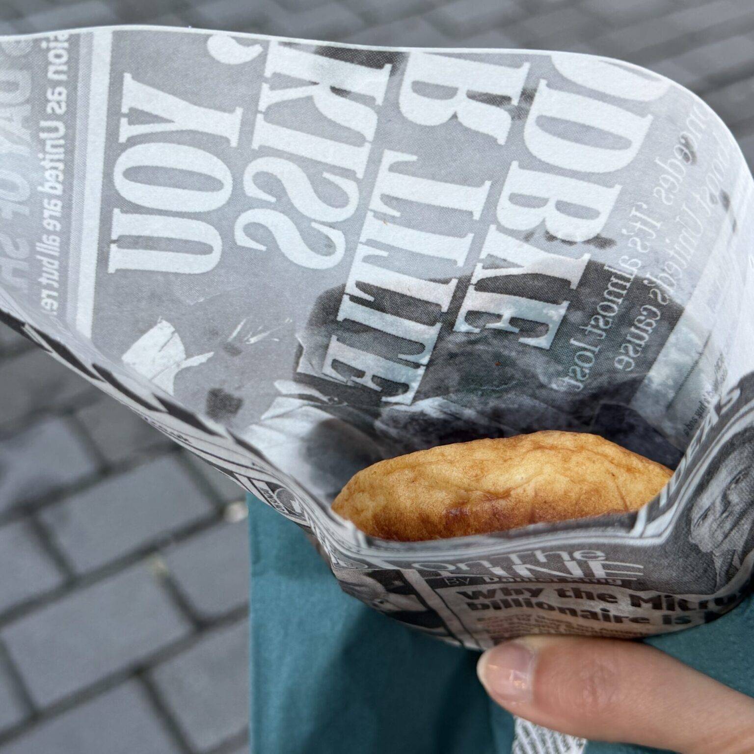 Eine Hand hält eine Fischfrikadelle, die in Zeitungspapier gewickelt ist, vor einem gepflasterten Boden.