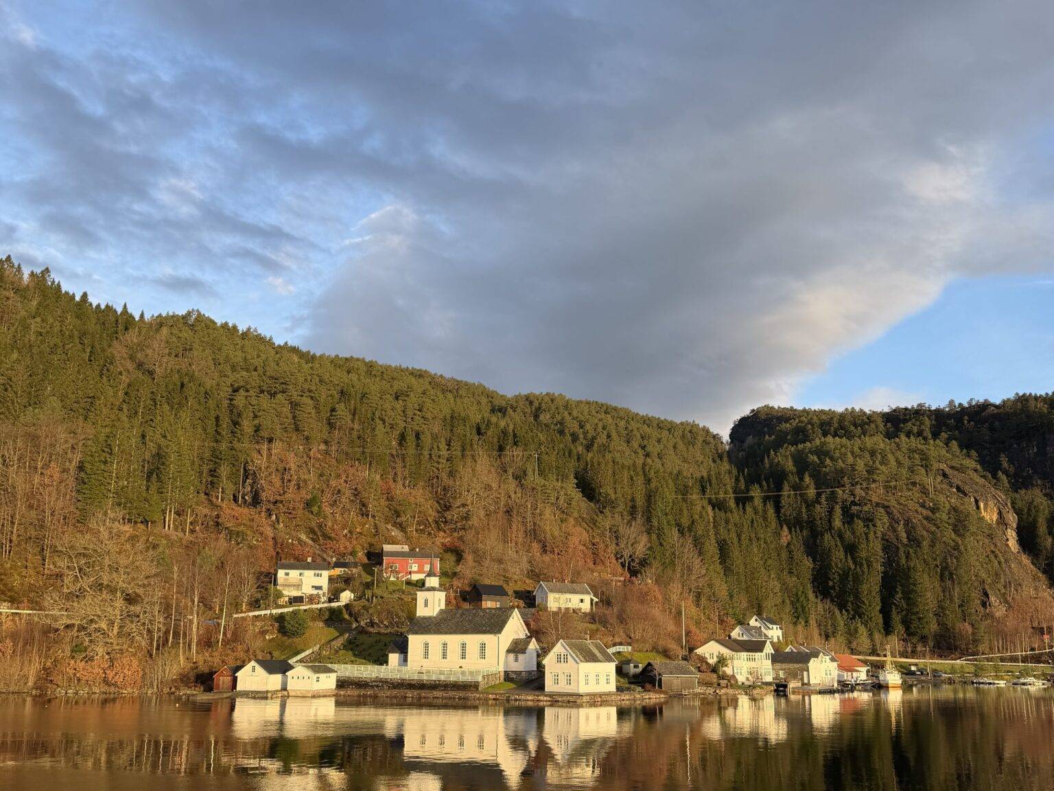 Mehrere Häuser am Wasser eines Fjords, umgeben von Wald und Hügeln, warmes Sonnenlicht auf den Fassaden.