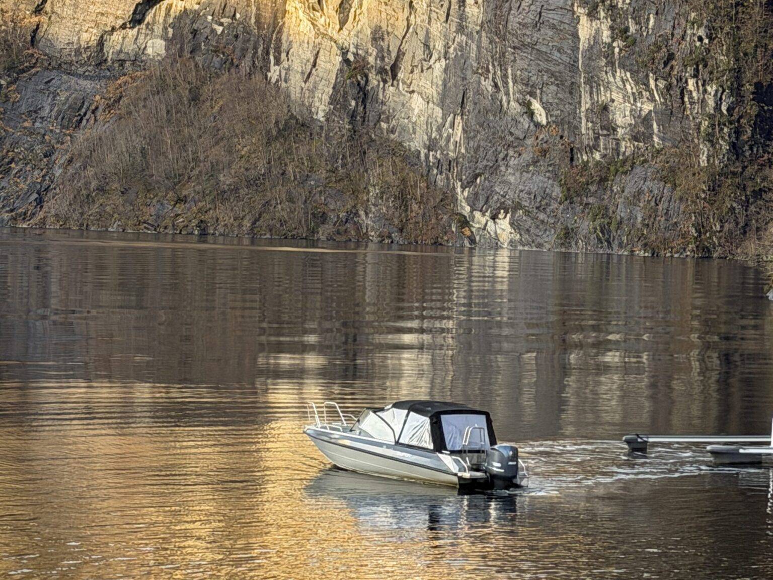 Ruhiger Fjord mit steiler Felswand in goldenem Licht, ein kleines Boot fährt durch das Wasser.