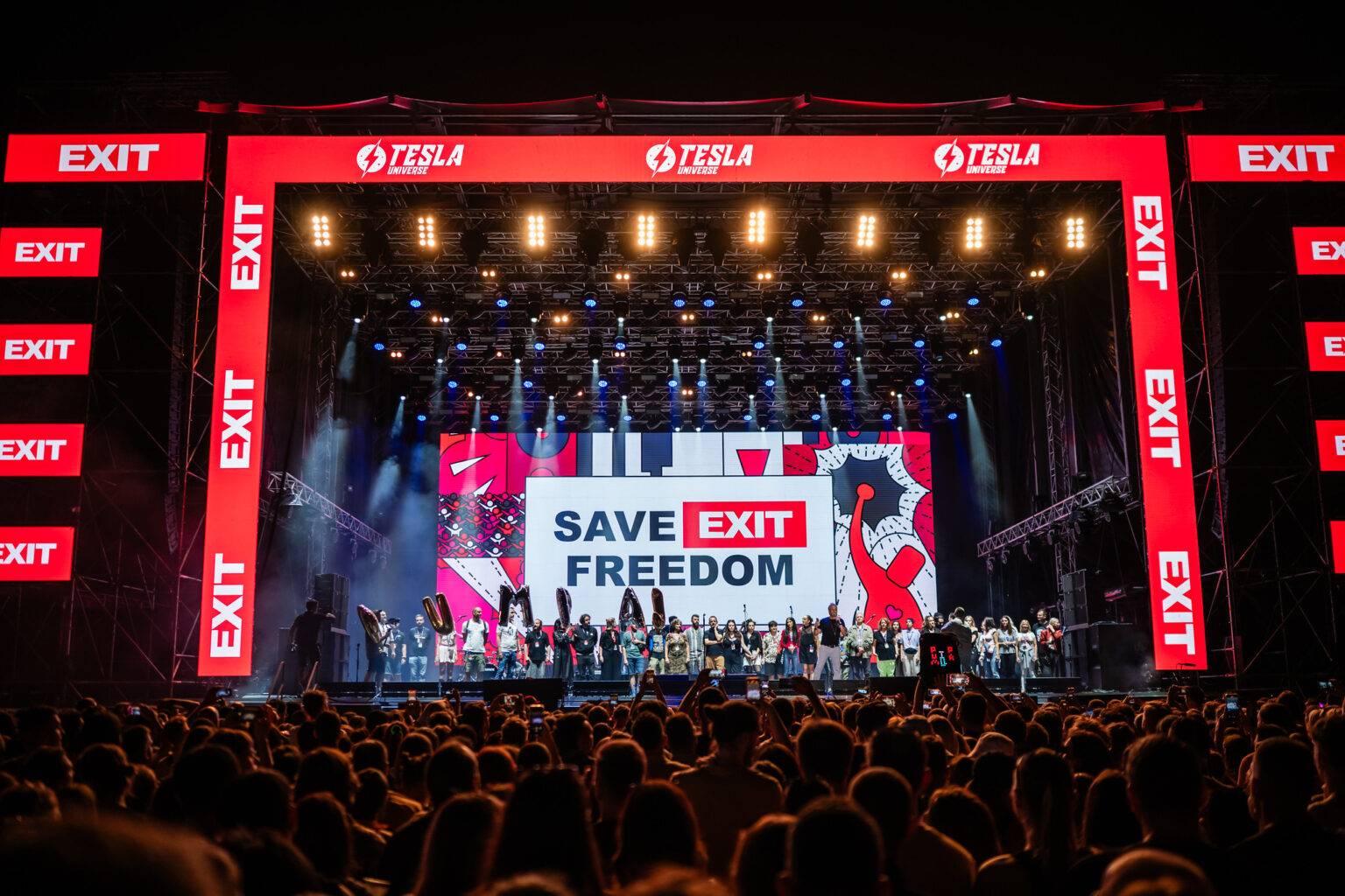 Bühne mit Menschen und einem großen Banner, auf dem "SAVE EXIT FREEDOM" steht, umgeben von leuchtenden Lichtern.