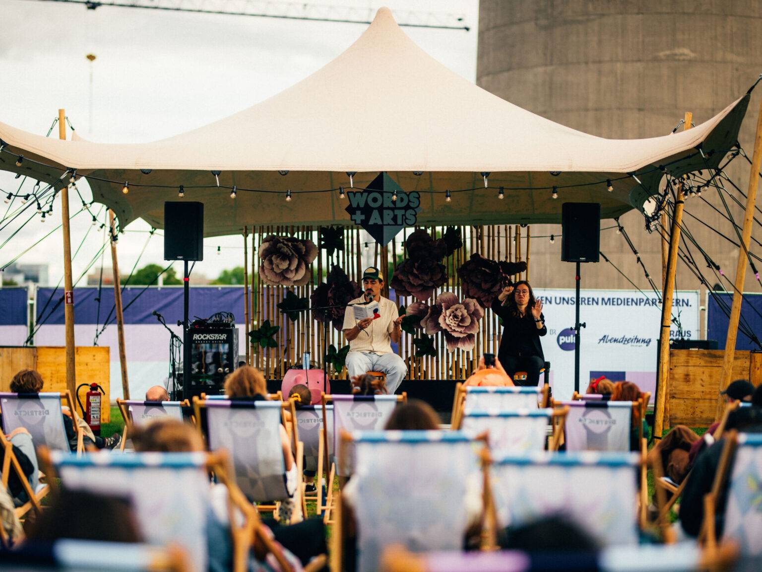 Bühne mit Zelt, ein Redner und eine Dolmetscherin mit Publikum in Liegestühlen im Freien.