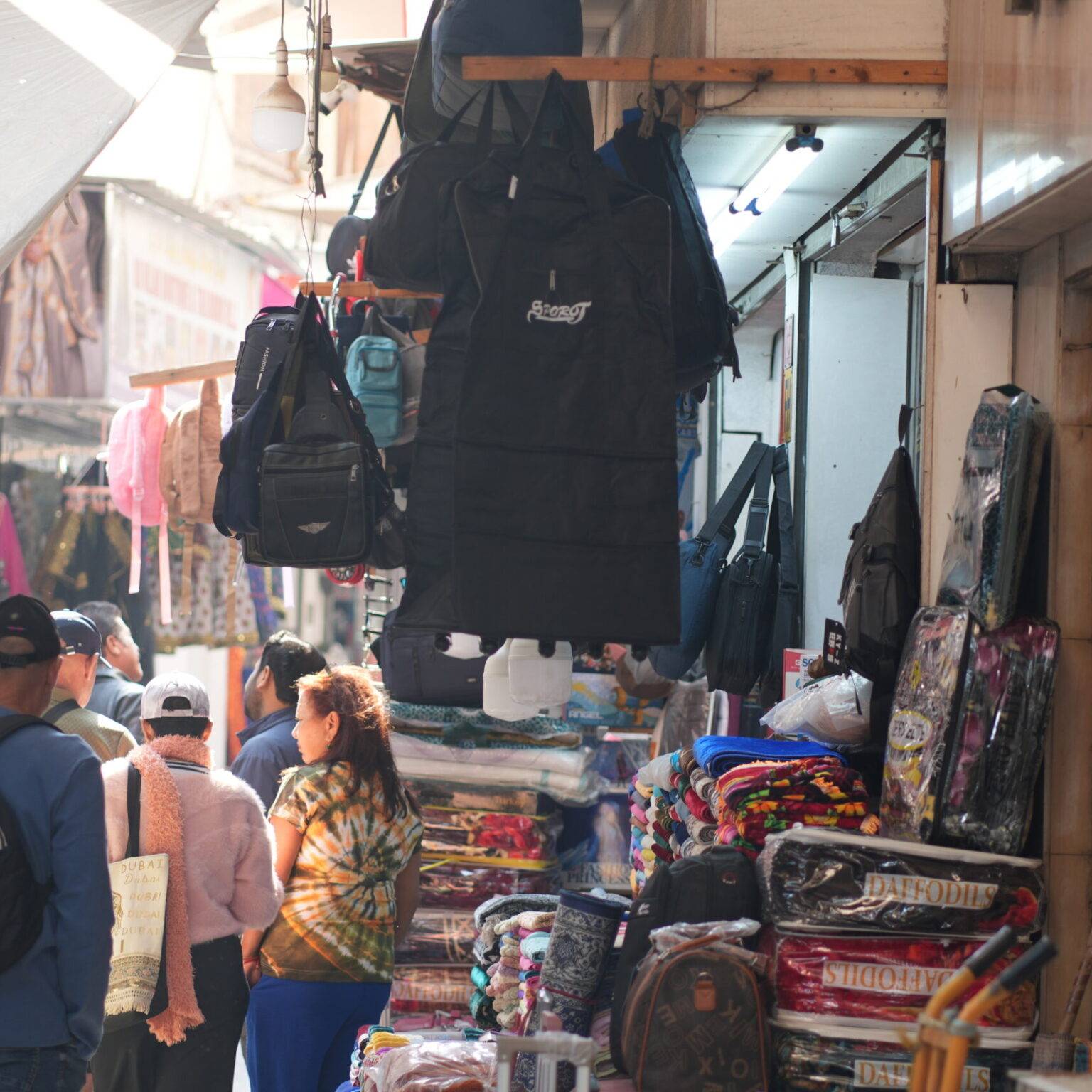 Belebte Gasse im Bab Al Bahrain Souk in Manama mit Händlern, Textilien und hängenden Taschen unter einem überdachten Marktgang.