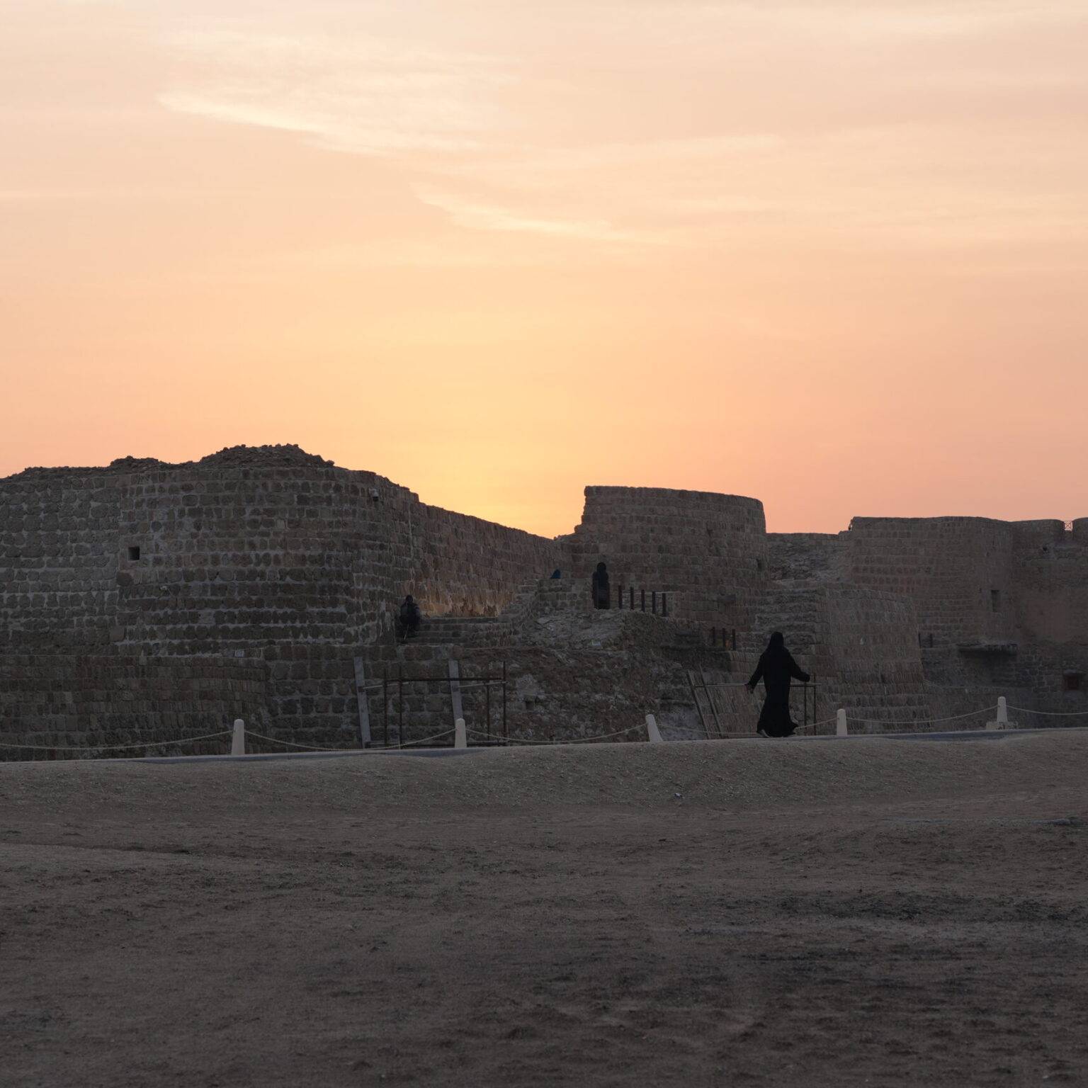 Ruinen silhouettiert gegen einen Sonnenuntergang am Horizont, mit einem sandigen Boden im Vordergrund.