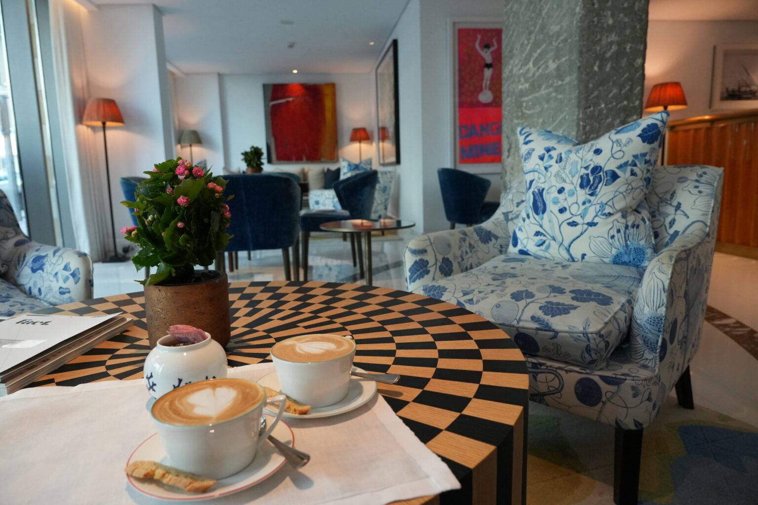 Zwei Tassen Kaffee auf einem Tisch mit Schachbrettmuster, umgeben von stilvollen Stühlen und einer Pflanze.