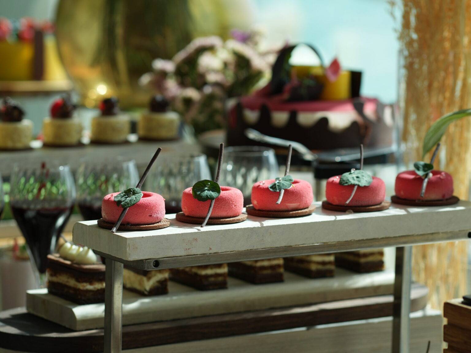 Vier rosa Cupcakes mit grünen Blättern als Deko auf einem Tablett vor weiteren Süßspeisen im Hintergrund.
