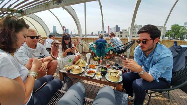 Auf der Dachterrasse des Aloha Bar sitzen junge Menschen um einen kleinen Tisch, der mit Speisen gedeckt ist, einige Gäste blicken vertieft in ihr Handy.