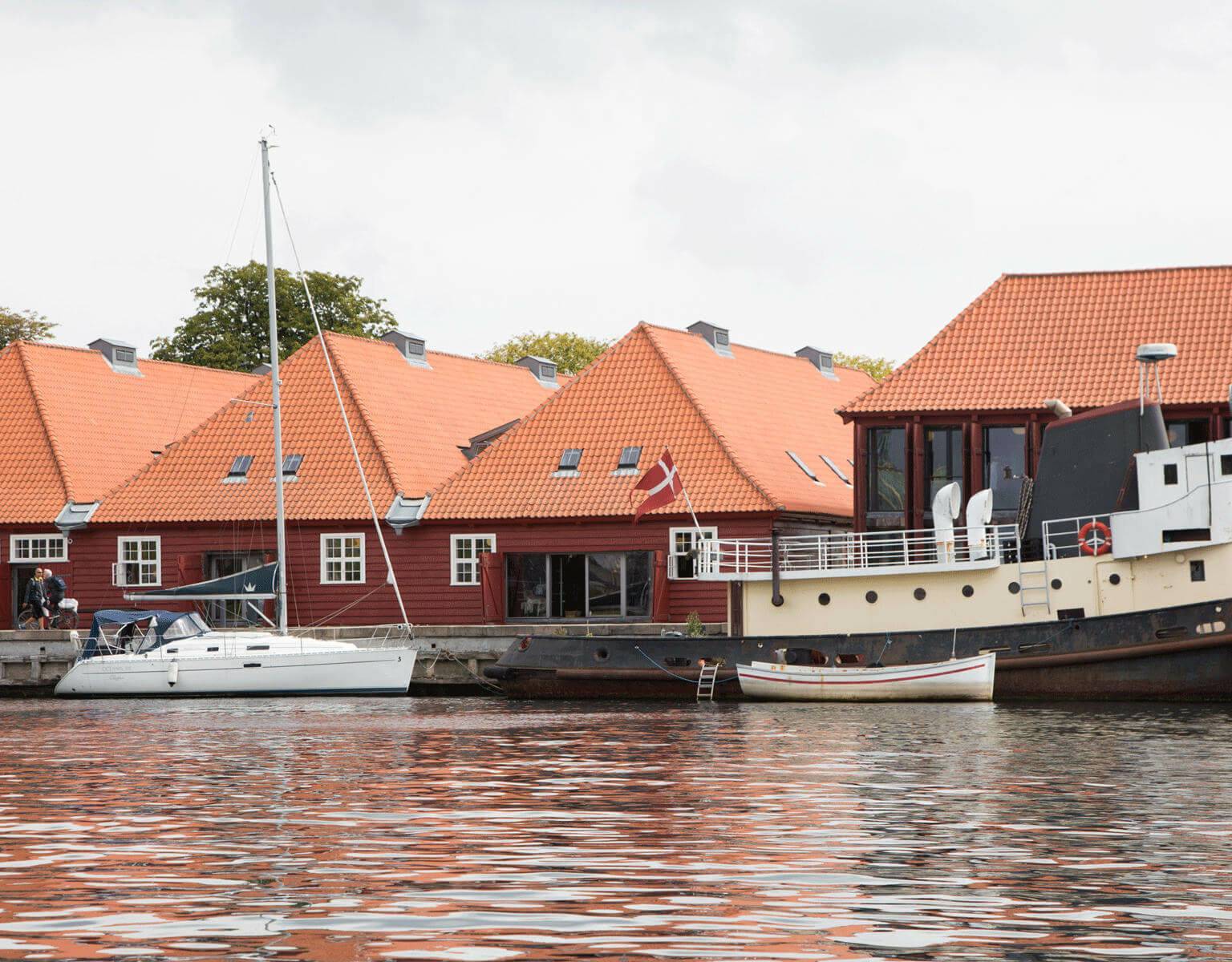 Ein Segelschiff und ein Schiff haben vor einem Ufer in Kopenhagen geankert, im Hintergrund stehen rote Holzhäuschen mit weißen Fenstern.