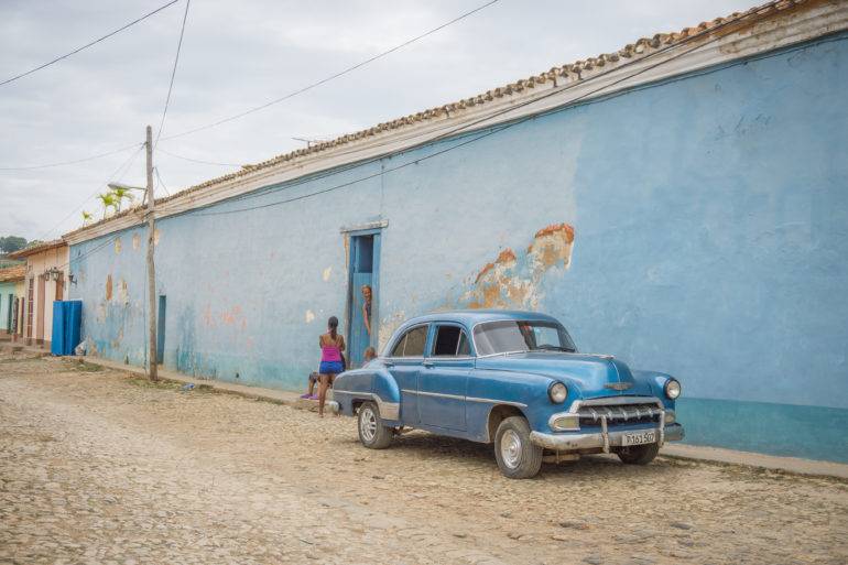 Auf einer Straße in Trinidad auf Kuba steht ein blauer Oldtimer vor einer blauen Hausfassade aus dessen blauer Tür ein Mann herausblickt und mit einem Mädchen auf der Straße redet.