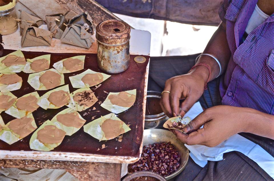 Auf dem Chowri Bazaar in Delhi füllt eine einheimische Frau im Sitzen Paan mit Limettenpaste und Betelnuss und hat diese auf einem Tablett vor ihr angerichtet.