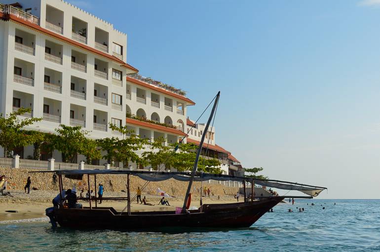 An der Küste einer Hotelanlage in Sansibar hat ein Dhow angelegt, im Hintergrund tummeln sich Strandgäste.