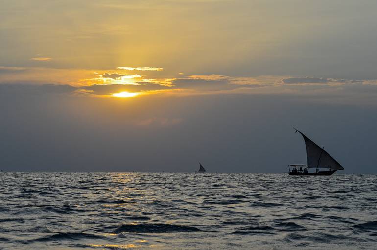 Auf dem Meer in Tansania verschwindet die Sonne hinter den Wolken während kleine Dhows mit Segeln friedlich dahin schippern.