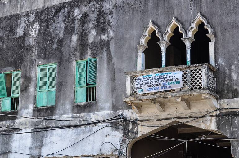 Eine heruntergekommene Hausfassade in Sansibar lässt indischen Einfluss vermuten, die grünen Fensterläden sind zum Teil rausgebrochen und viele Kabel laufen wirr der Hausfront entlang.