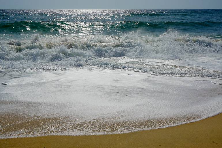 In der Sonne glitzert das Wasser des Meeres während eine Welle am gelben Sandstrand an der Algarve bricht.