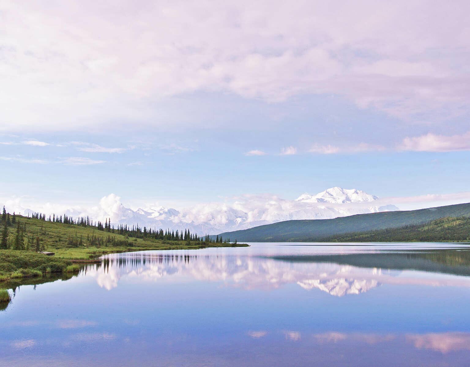 In Alaska am Yukon im Denali Nationalpark, USA liegt ein Bergsee vor den schneebedeckten Bergen zwischen bewaldeten Wiesenhügeln.