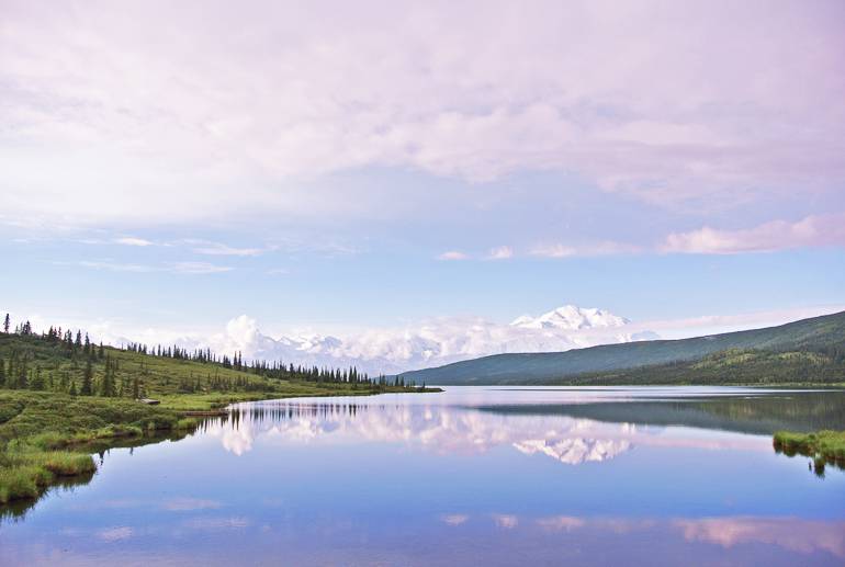In Alaska am Yukon im Denali Nationalpark, USA liegt ein Bergsee vor den schneebedeckten Bergen zwischen bewaldeten Wiesenhügeln.