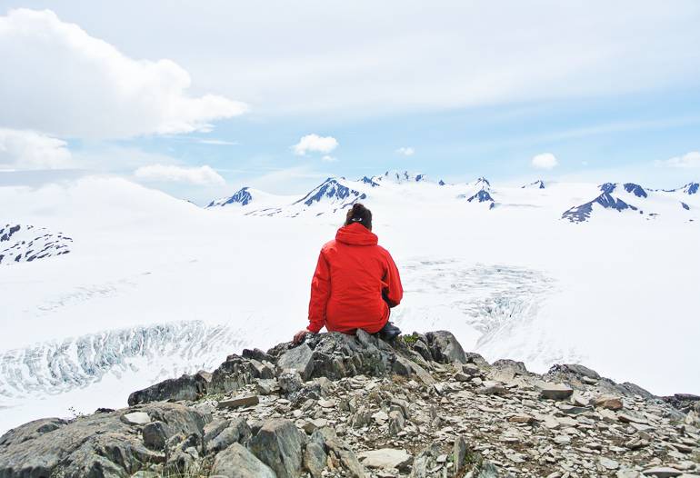 Eine Wanderin mit roter Jacke und schwarzer Mütze sitzt auf einem steinigen Felsvorsprung und betrachtet die schneebedeckte Gletscherlandschaft von Harding Icefield, Alaska.