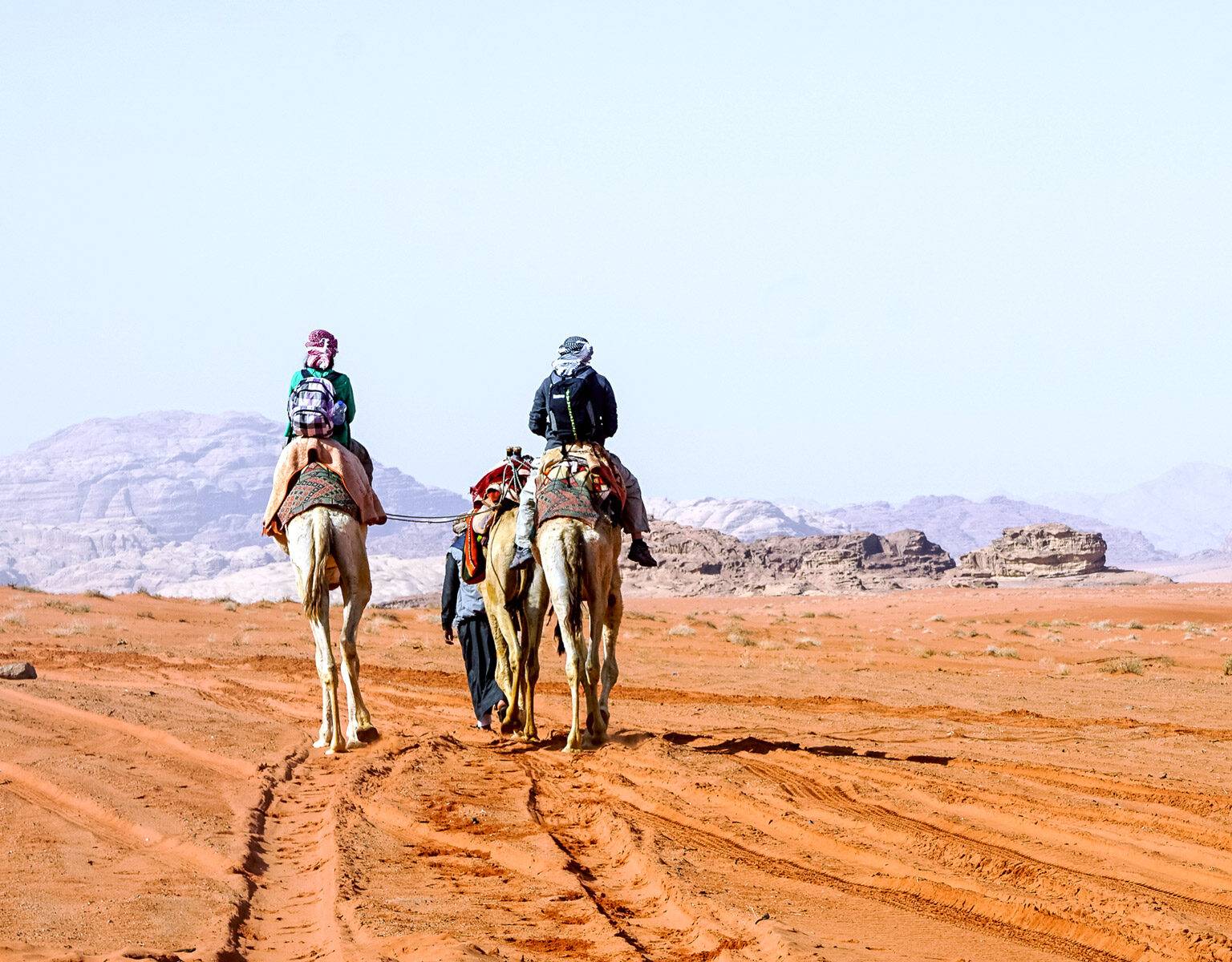 Zwei Touristen mit Gepäck werden auf Kamelen von einem Einheimischen durch die Wüste Wadi Rum in Jordanien geführt.