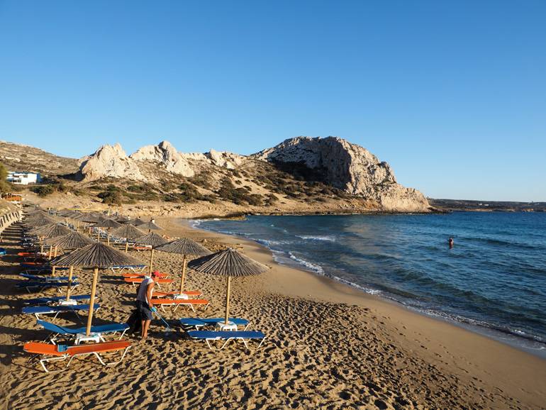An Karpathos Strand Ágios Theódoros reihen sich Strohschirme und blau- und orangefarbene Liegen entlang des Sandstrandes, im Hintergrund ragt eine Felsformation aus dem dunkelblauen Meer.