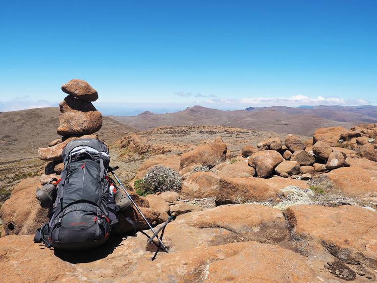 Südafrika Drakensberge: Ein Rucksack steht auf dem Boden, im Hintergrund ein Turm aus Steinen.