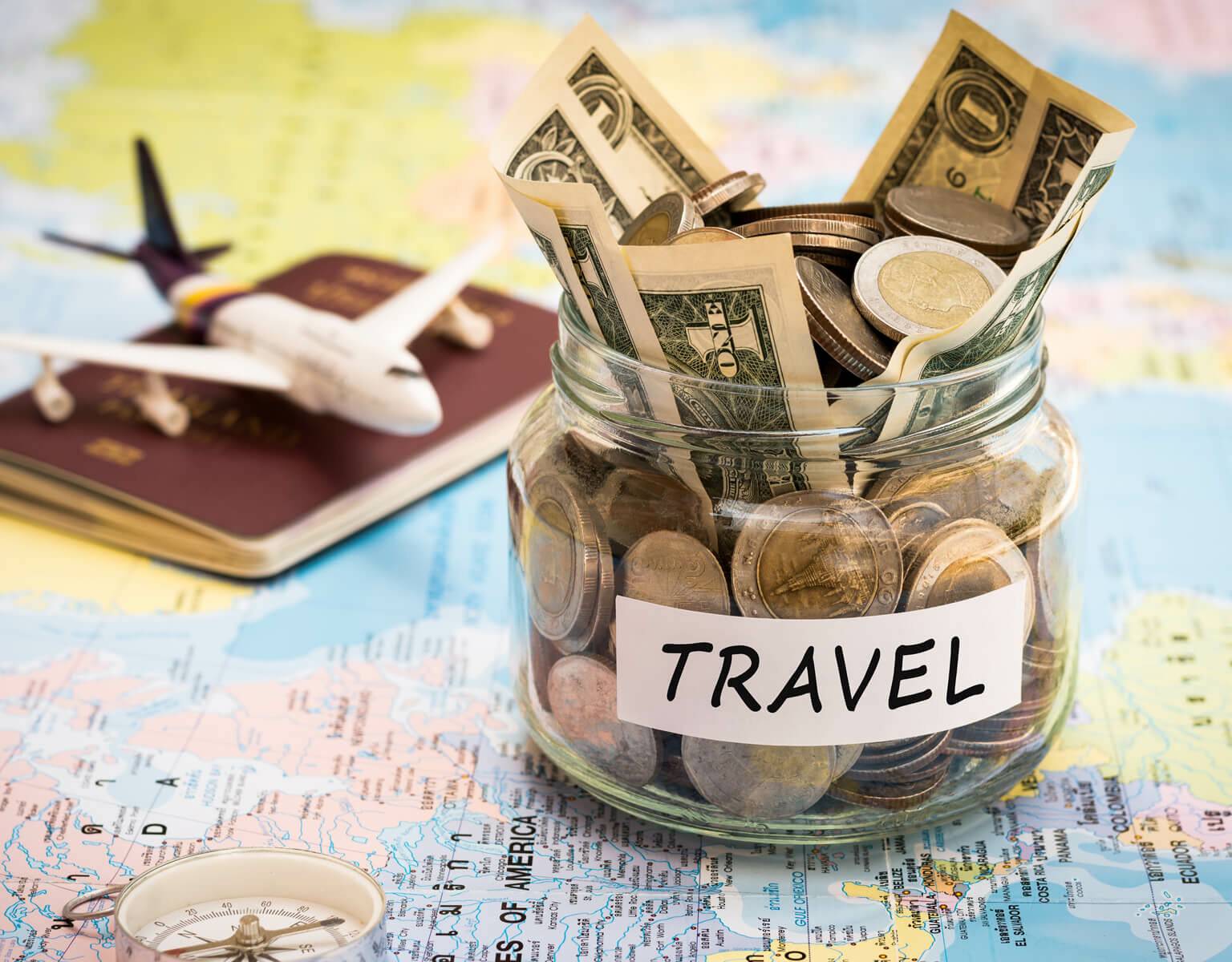 Ein Weckglas mit Scheinen und Münzen gefüllt steht auf einer Weltkarte, dahinter Reisepass und Mini-Flugzeug.