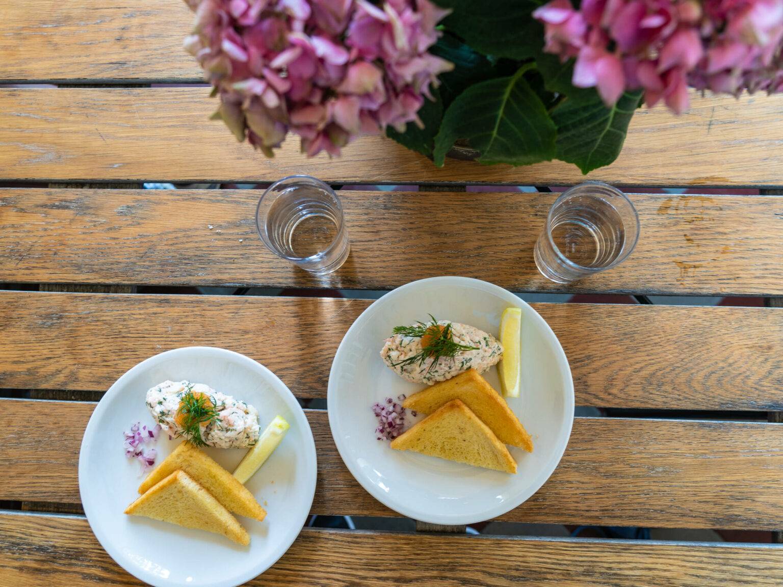 Auf einem Holztisch stehen zwei Teller mit dem typisch schwedischen Gericht Toast Skagen, ein Garnelensalat mit Sauerrahm und Dill mit Toastscheiben.