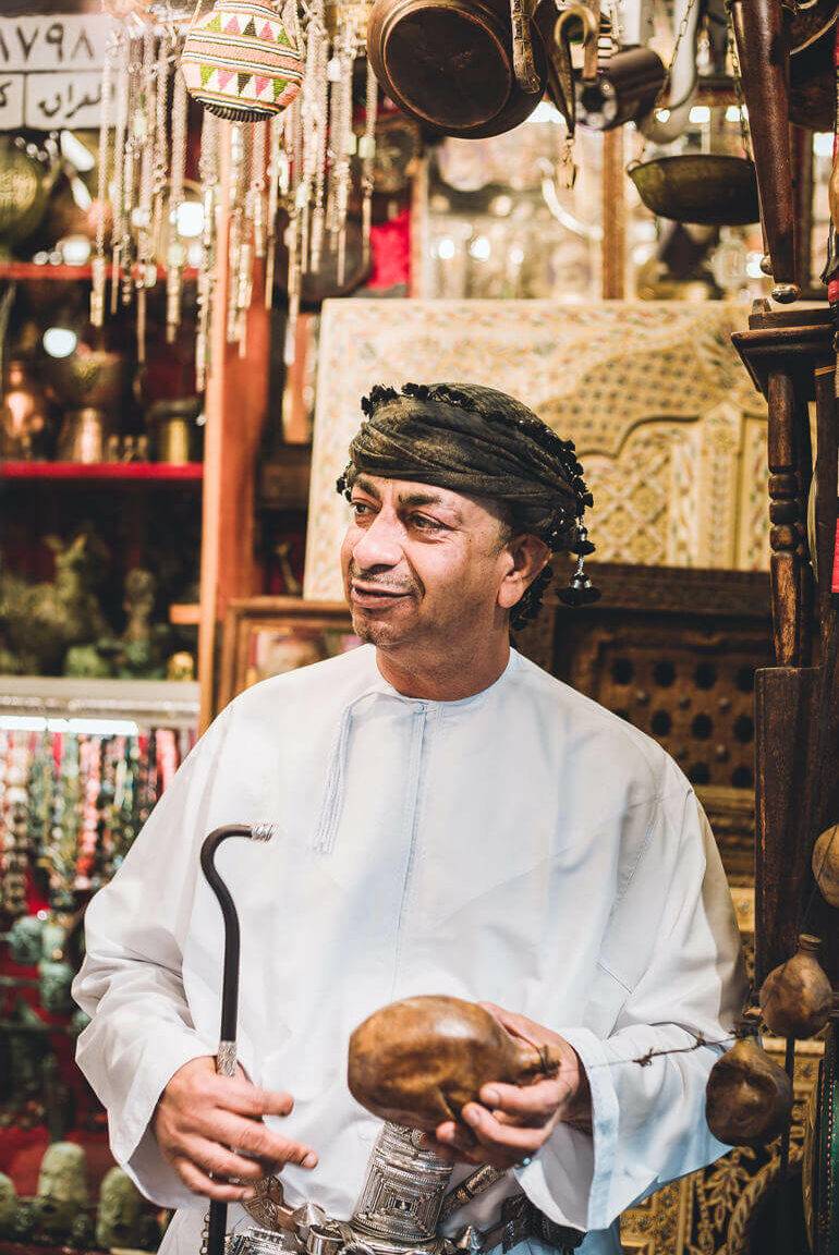 Roadtrip durch den Oman: Ein Mann mit Kopfbedeckung in einem Basar Geschäft hält einen Stock in der Hand.