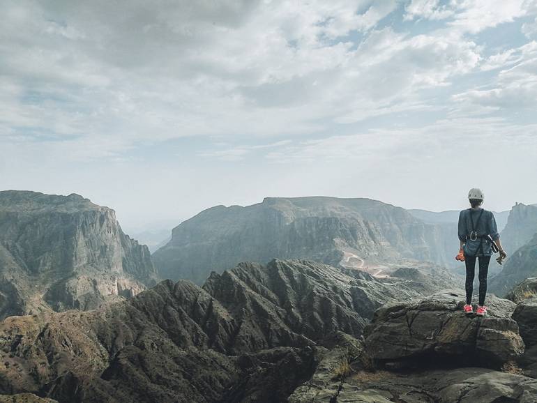 Roadtrip durch den Oman: Eine Frau von hinten, gekleidet in Kletterausrüstung, blickt auf die bergige Landschaft.