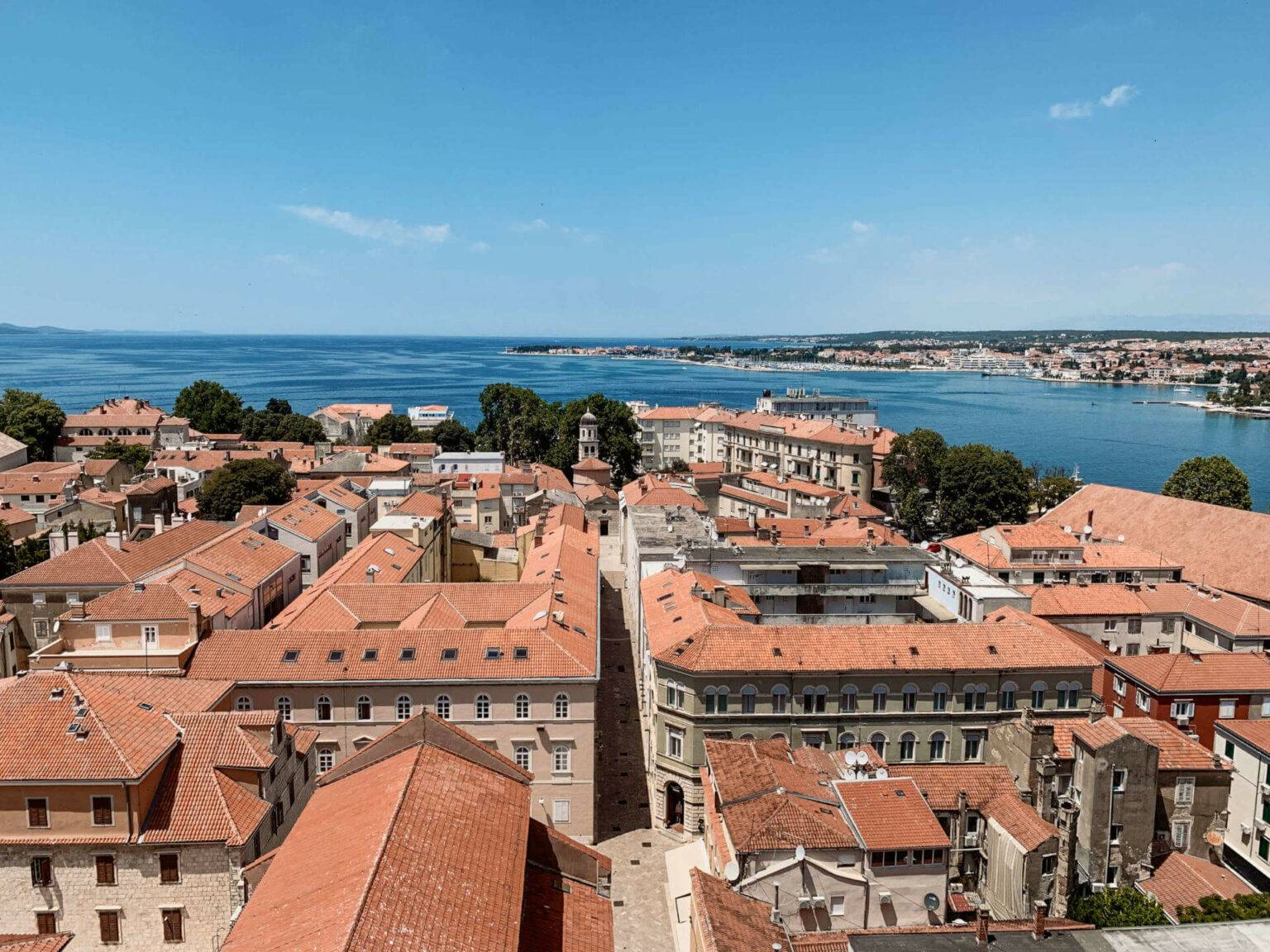 Blick vom Kirchturm über die roten Dächer der Altstadt von Zadar in Kroatien, in der Ferne das Meer.