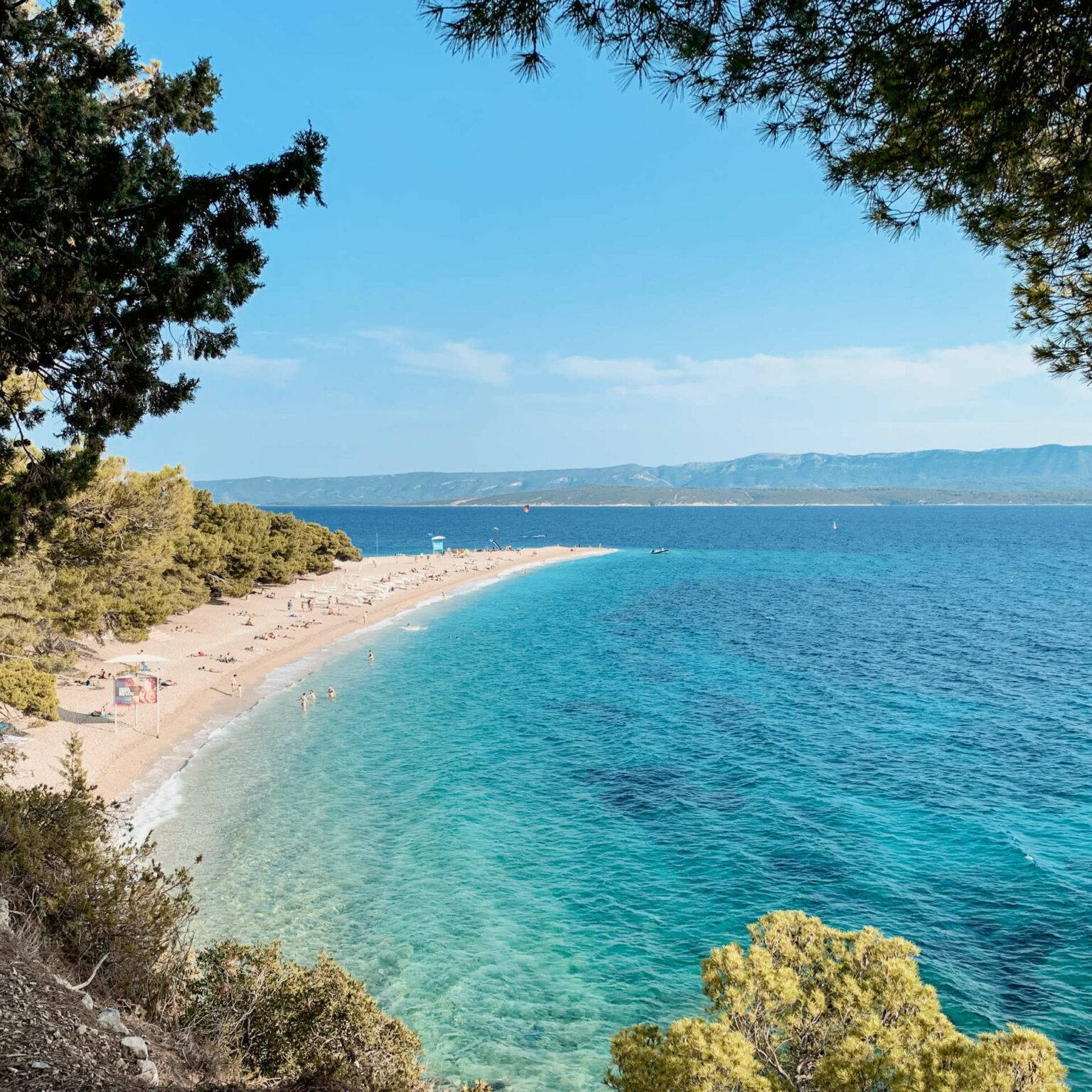 Blick durch Bäume auf den Sandstrand "Goldenes Horn" in Kroatien, der Reisende mit seinem türkisblauen Meer zum Baden einlädt.