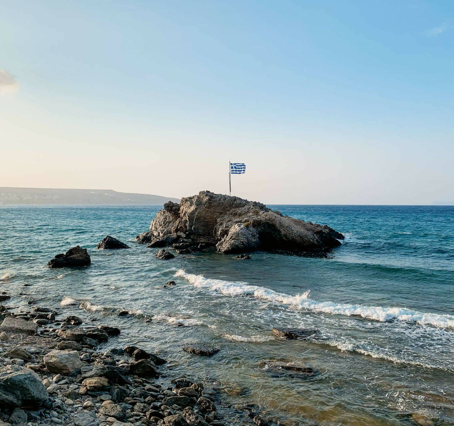 Leichter Wellengang an einem Steinstrand, aus dem türkisblauen Wasser erhebt sich ein Felsen mit Griechenlands Flagge darauf.
