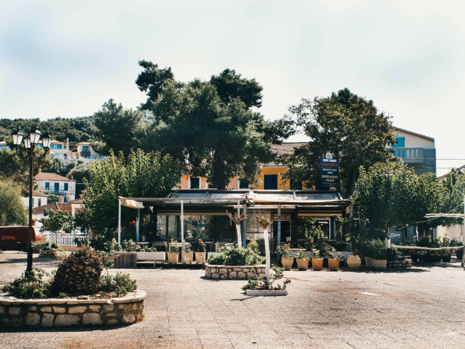 Ein gepflasterter Platz auf der Insel Meganisi, ein gelbes Haus ist hinter vielen grünen Bäumen zu erkennen.