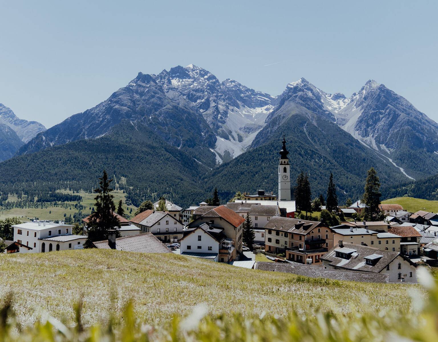 Blick über eine Wiese hinein in ein Dorf mit Kirchturmspitze, dahinter Bergpanorama, in Ftan in der Schweiz.